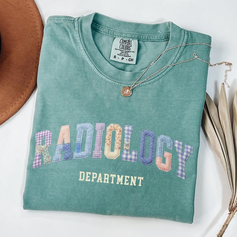 May include: A teal green t-shirt with the word "RADIOLOGY" in patchwork letters and "DEPARTMENT" printed below. A silver necklace with a pendant rests on the shirt. The shirt is folded and placed on a white surface.