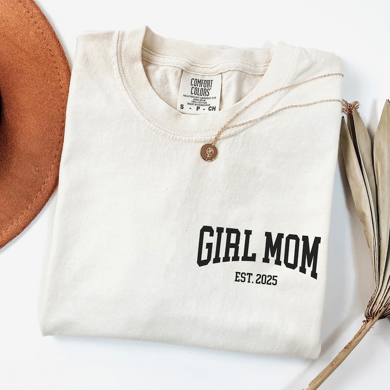 Op de afbeelding: Cr&egrave;mekleurig T-shirt met de tekst "GIRL MOM EST. 2025" in het zwart gedrukt. Een gouden ketting met een hanger ligt op het shirt. Een bruine hoed en gedroogde plantenstengels zijn op de achtergrond.