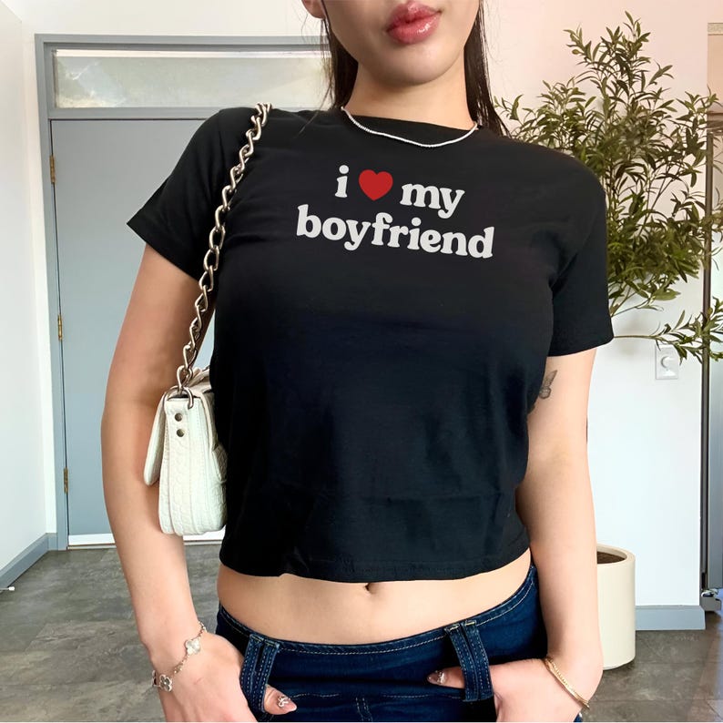 May include: A black cropped t-shirt with the text "i &hearts; my boyfriend" in white lettering. The shirt is being worn by a person with a white purse and blue jeans.