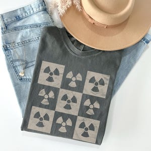 May include: A dark gray t-shirt with a checkered pattern of nine beige radiation symbols.