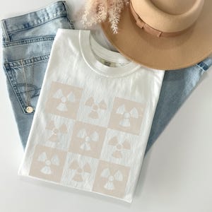 May include: White t-shirt with a beige pattern of repeating radiation symbols.
