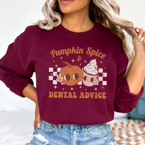 May include: Burgundy sweatshirt with a graphic of a pumpkin and a coffee cup with whipped cream. The text reads "Pumpkin Spice & Dental Advice".