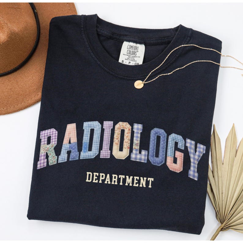 May include: Navy blue t-shirt with the word "RADIOLOGY" in patchwork letters and "DEPARTMENT" underneath. A gold necklace and a brown hat are also visible. The shirt is from Comfort Colors.