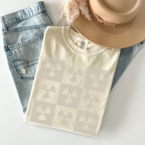 May include: A cream-colored t-shirt with a pattern of nine light gray radiation symbols. The shirt is laid out on a white surface with a pair of blue jeans and a straw hat.