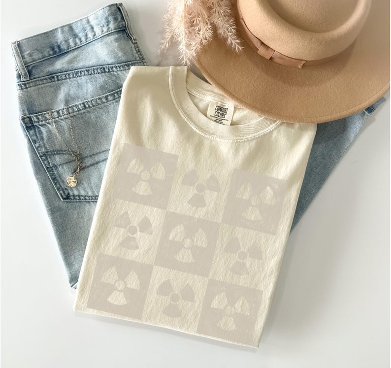 May include: A cream-colored t-shirt with a pattern of nine light gray radiation symbols. The shirt is laid out on a white surface with a pair of blue jeans and a straw hat.