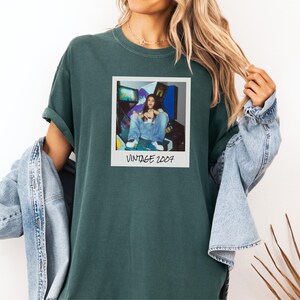 Pode incluir: T-shirt verde escuro com uma imagem em estilo polaroid mostrando uma pessoa de macac&atilde;o jeans em frente a jogos de arcade. A polaroid tem o texto "VINTAGE 2007". A camisa tem mangas enroladas.