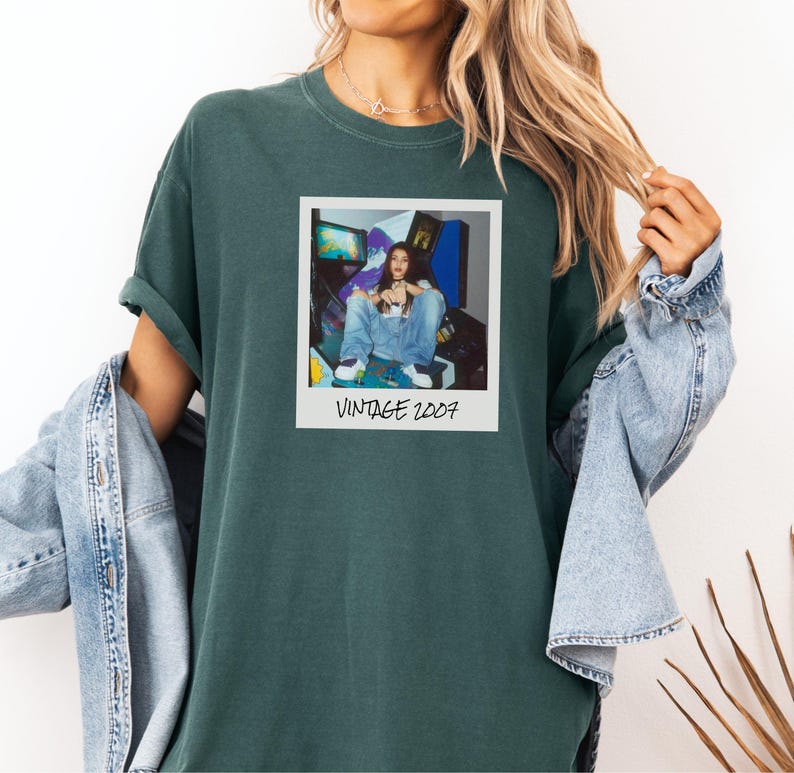 Pode incluir: T-shirt verde escuro com uma imagem em estilo polaroid mostrando uma pessoa de macac&atilde;o jeans em frente a jogos de arcade. A polaroid tem o texto "VINTAGE 2007". A camisa tem mangas enroladas.