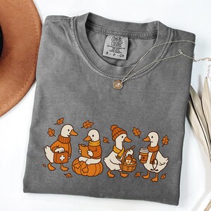 May include: A gray Comfort Colors t-shirt featuring a whimsical design of four ducks dressed in fall attire. The ducks are depicted with a pumpkin, a basket, and coffee cups, surrounded by autumn leaves. A silver necklace with a pendant is also visible.