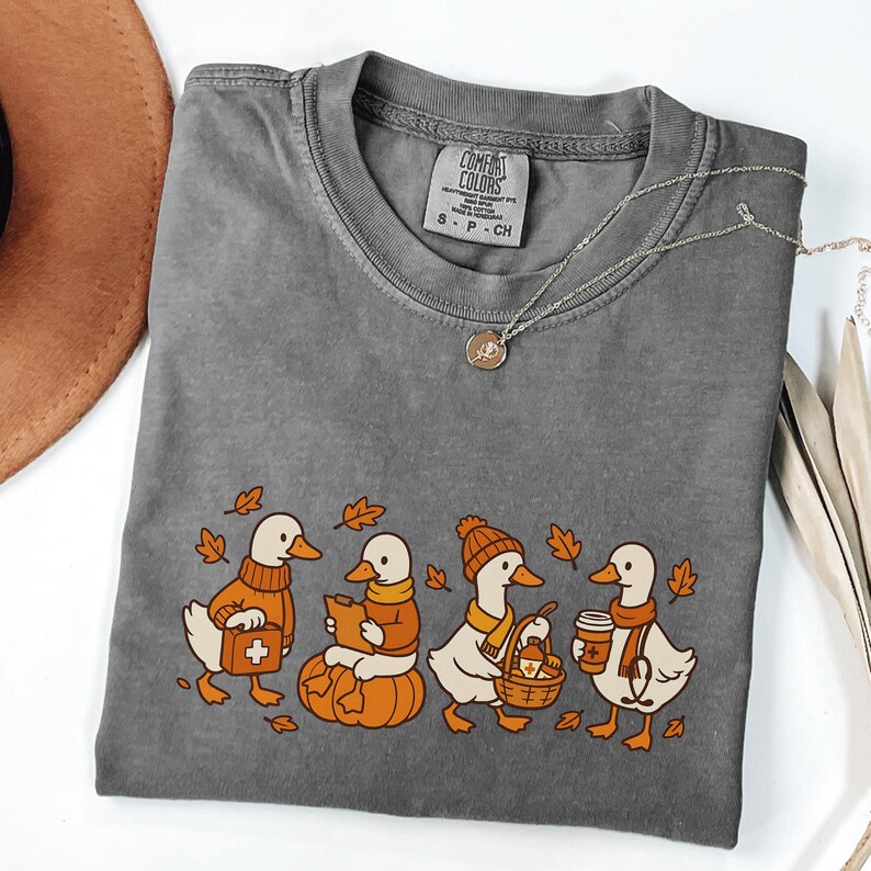May include: A gray Comfort Colors t-shirt featuring a whimsical design of four ducks dressed in fall attire. The ducks are depicted with a pumpkin, a basket, and coffee cups, surrounded by autumn leaves. A silver necklace with a pendant is also visible.