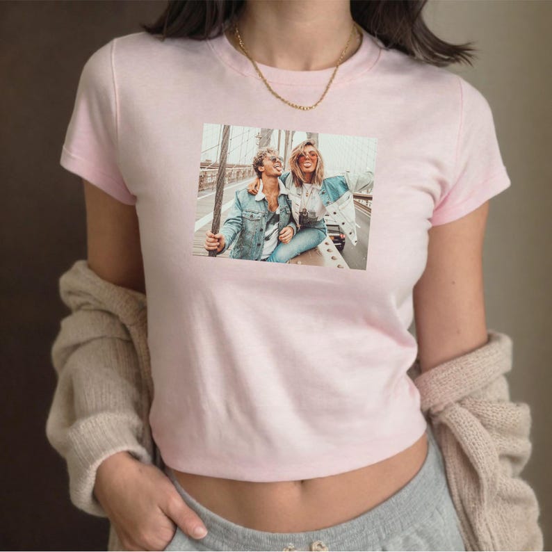 May include: A light pink crop top with a crew neck and short sleeves. The shirt features a rectangular graphic of two people on a bridge. A gold necklace is visible. A beige sweater is draped over the shoulders.