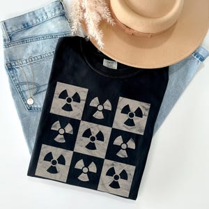 May include: Black t-shirt with a pattern of nine gray radiation symbols on a black background. The shirt is folded and lying on a blue denim background.