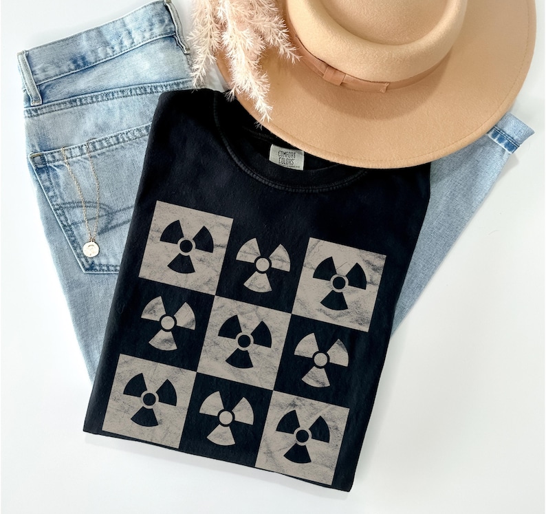 May include: Black t-shirt with a pattern of nine gray radiation symbols on a black background. The shirt is folded and lying on a blue denim background.