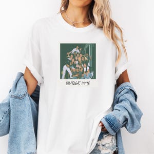 Pode incluir: Camiseta branca com uma fotografia em estilo vintage de um grupo de pessoas. A foto &eacute; quadrada e tem o texto "VINTAGE 1998" impresso abaixo. A camiseta tem as mangas dobradas.