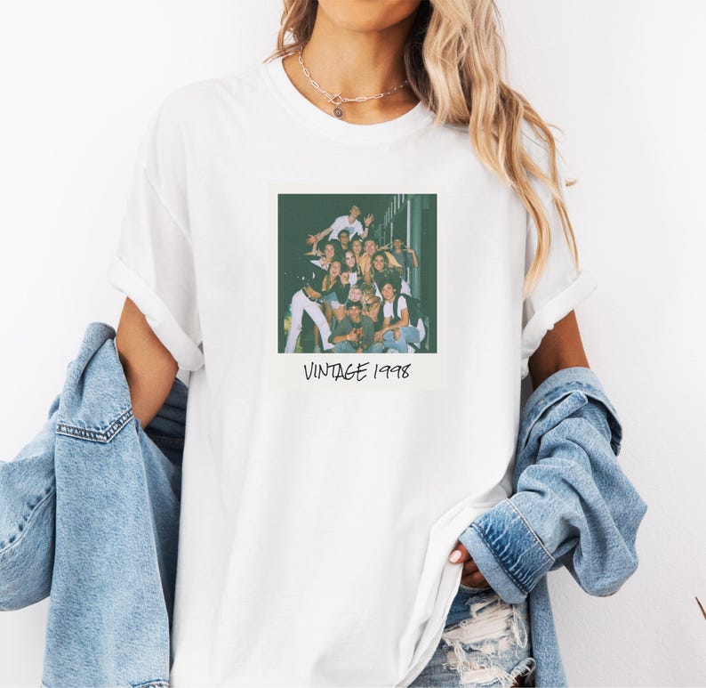 Pode incluir: Camiseta branca com uma fotografia em estilo vintage de um grupo de pessoas. A foto &eacute; quadrada e tem o texto "VINTAGE 1998" impresso abaixo. A camiseta tem as mangas dobradas.