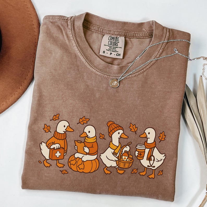 May include: Brown Comfort Colors t-shirt featuring a cartoon design of four ducks dressed in fall attire. The ducks are carrying items like a first aid kit, a book, a basket, and a coffee cup. A silver necklace with a pendant is also visible.