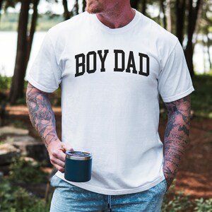 May include: White t-shirt with the text "BOY DAD" in bold black letters. The shirt is short-sleeved and appears to be made of cotton. A person is holding a dark blue insulated tumbler. The background is a blurred outdoor scene.