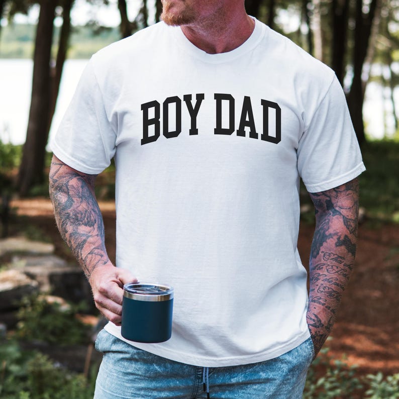 May include: White t-shirt with the text "BOY DAD" in bold black letters. The shirt is short-sleeved and appears to be made of cotton. A person is holding a dark blue insulated tumbler. The background is a blurred outdoor scene.