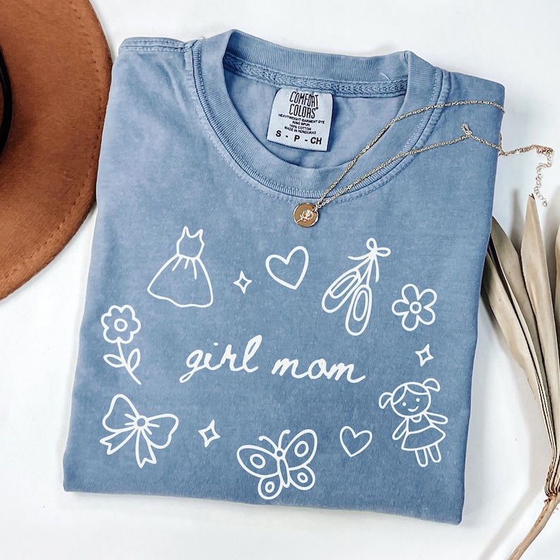May include: A blue Comfort Colors t-shirt with the words "girl mom" and various white illustrations, including a dress, hearts, ballet shoes, and a butterfly. A gold necklace with a pendant rests on the shirt.