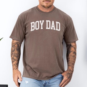 May include: A brown t-shirt with the text "BOY DAD" in white block letters. The shirt has short sleeves and a crew neck. The person wearing the shirt has tattoos on both arms.
