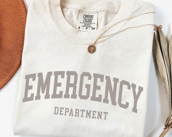 May include: A beige t-shirt with the text "EMERGENCY DEPARTMENT" in gray letters. The shirt is folded and has a gold necklace with a small pendant on top.