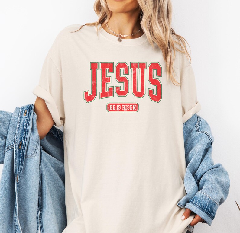 Pu&ograve; includere: Una t-shirt color crema con una grafica vintage in rosso e verde che dice "JESUS" con le parole "HE IS RISEN" in un font pi&ugrave; piccolo sotto.