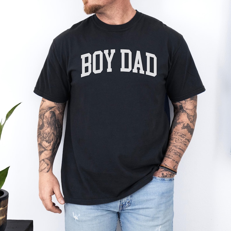 May include: Black t-shirt featuring the white text "BOY DAD" in an arched design. The t-shirt has a crew neck. The person wearing the shirt has tattoos on their arms and is wearing blue jeans.