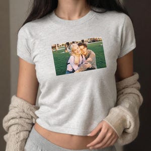 May include: A light gray t-shirt featuring a rectangular image of a couple embracing on a grassy field. The couple is smiling and appears to be happy. The t-shirt is short-sleeved and made of a soft material.