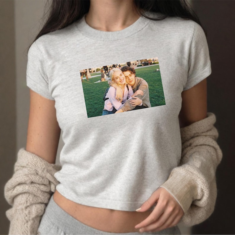 May include: A light gray t-shirt featuring a rectangular image of a couple embracing on a grassy field. The couple is smiling and appears to be happy. The t-shirt is short-sleeved and made of a soft material.