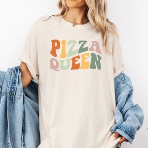 May include: A light beige t-shirt with the text "PIZZA QUEEN" in colorful retro-style lettering.