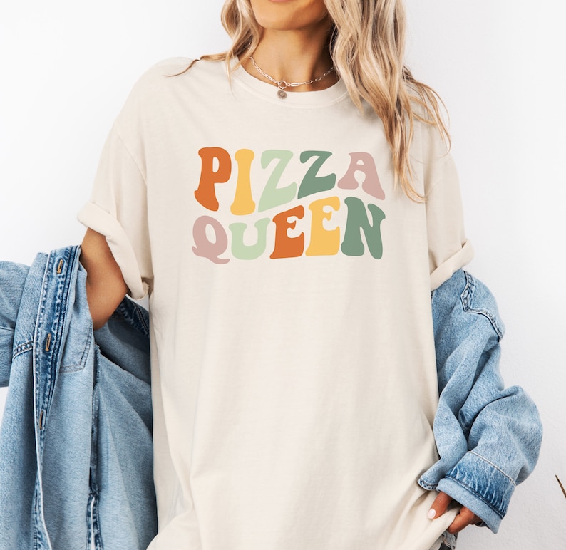 May include: A light beige t-shirt with the text "PIZZA QUEEN" in colorful retro-style lettering.