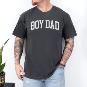 May include: Dark grey V-neck t-shirt with the white text "BOY DAD" in a collegiate font. The shirt is made of a soft material and has short sleeves. The shirt is worn by a person.