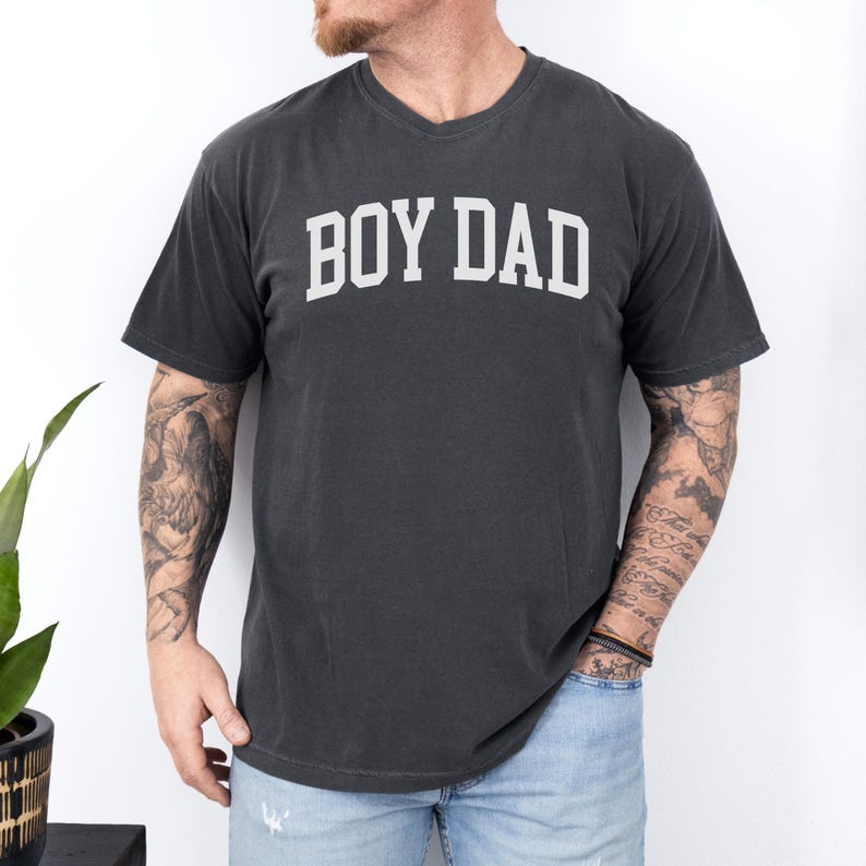 May include: Dark grey V-neck t-shirt with the white text "BOY DAD" in a collegiate font. The shirt is made of a soft material and has short sleeves. The shirt is worn by a person.