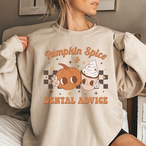 May include: A beige sweatshirt with a graphic design of a pumpkin and a cup of coffee. The text on the sweatshirt reads "Pumpkin Spice & Dental Advice".