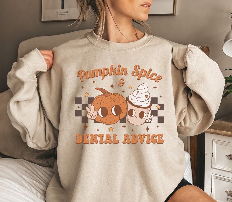 May include: A beige sweatshirt with a graphic design of a pumpkin and a cup of coffee. The text on the sweatshirt reads "Pumpkin Spice & Dental Advice".
