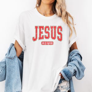 Pu&ograve; includere: Maglietta bianca con testo rosso che dice "JESUS" in un font universitario vintage. Sotto il testo c'&egrave; un rettangolo rosso pi&ugrave; piccolo con il testo "HE IS RISEN".