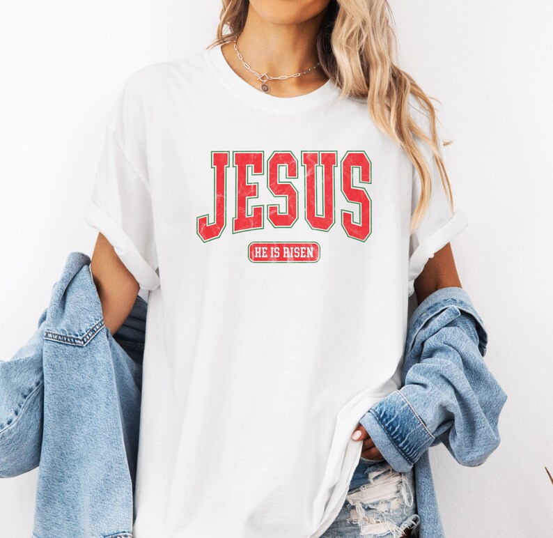 Pu&ograve; includere: Maglietta bianca con testo rosso che dice "JESUS" in un font universitario vintage. Sotto il testo c'&egrave; un rettangolo rosso pi&ugrave; piccolo con il testo "HE IS RISEN".