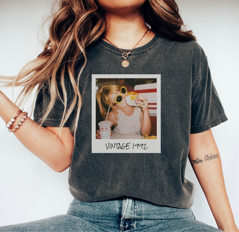 Pode incluir: T-shirt cinza escuro com uma imagem vintage em estilo Polaroid de uma crian&ccedil;a usando &oacute;culos de sol e comendo um hamb&uacute;rguer. A imagem est&aacute; rotulada "VINTAGE 1992" em branco. A camisa tem uma apar&ecirc;ncia ligeiramente desbotada.