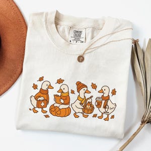 May include: Cream-colored t-shirt featuring a line of cartoon ducks in autumn attire. The ducks are wearing sweaters, scarves, and hats, and carrying items like a first aid kit, pumpkin, basket, and coffee. The shirt has the text "COMFORT COLORS".