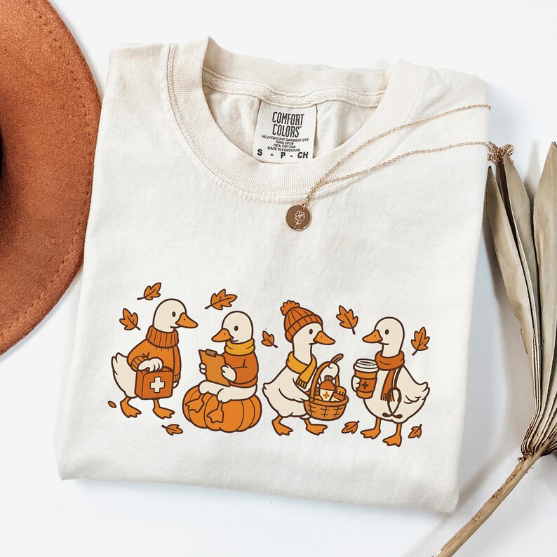 May include: Cream-colored t-shirt featuring a line of cartoon ducks in autumn attire. The ducks are wearing sweaters, scarves, and hats, and carrying items like a first aid kit, pumpkin, basket, and coffee. The shirt has the text "COMFORT COLORS".