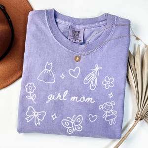 May include: A lilac-colored t-shirt with the words "girl mom" in cursive, surrounded by white illustrations of a dress, hearts, ballet shoes, flowers, a bow, a butterfly, and a girl. A gold necklace with a pendant is on the shirt.
