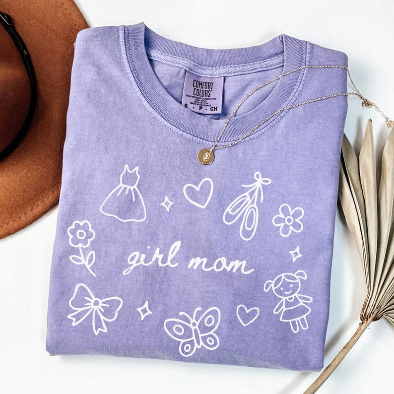May include: A lilac-colored t-shirt with the words "girl mom" in cursive, surrounded by white illustrations of a dress, hearts, ballet shoes, flowers, a bow, a butterfly, and a girl. A gold necklace with a pendant is on the shirt.