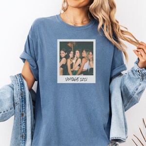 Pode incluir: Uma camiseta azul com uma fotografia em estilo vintage com o texto "VINTAGE 2021". A fotografia mostra um grupo de pessoas. A camisa tem mangas enroladas e &eacute; usada com uma jaqueta jeans.