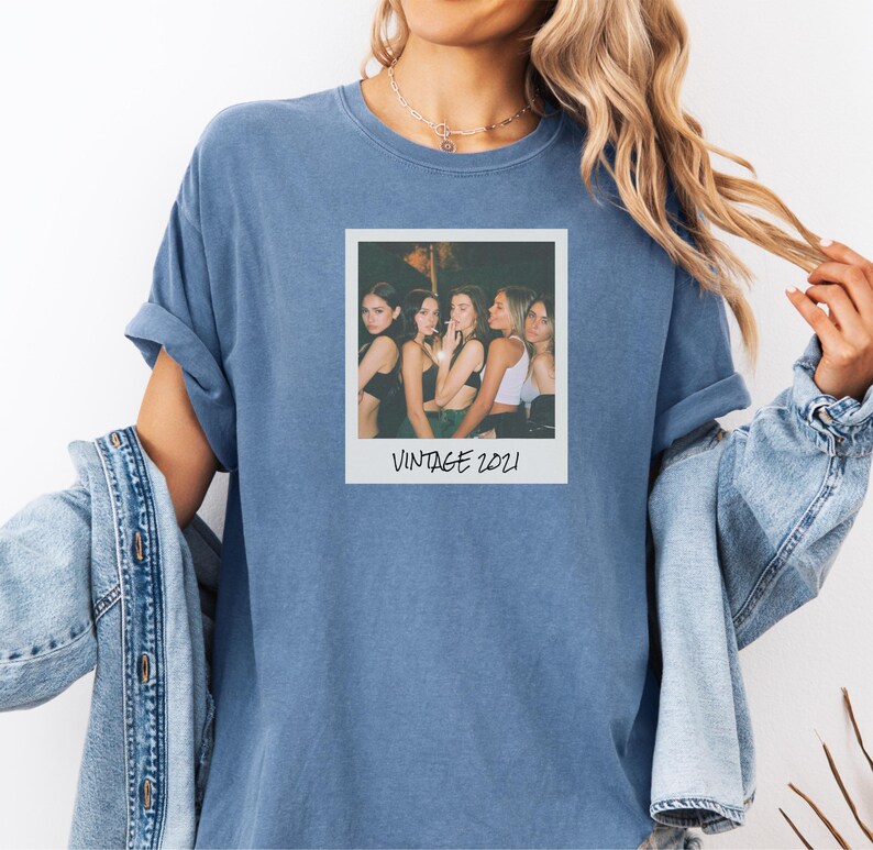 Pode incluir: Uma camiseta azul com uma fotografia em estilo vintage com o texto "VINTAGE 2021". A fotografia mostra um grupo de pessoas. A camisa tem mangas enroladas e &eacute; usada com uma jaqueta jeans.