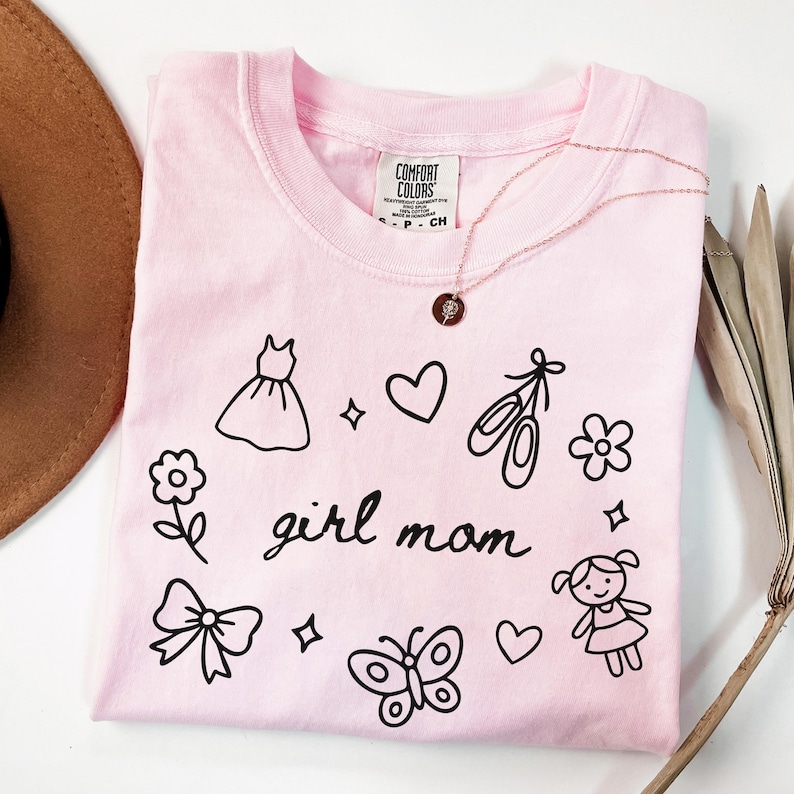 May include: A light pink t-shirt with the words "girl mom" in a handwritten font, surrounded by black line drawings of a dress, hearts, ballet shoes, a flower, a bow, a butterfly, and a girl. A gold necklace is draped on the shirt.