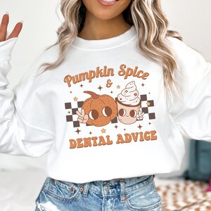 May include: White sweatshirt with a graphic of a pumpkin and a coffee cup with whipped cream. The text "Pumpkin Spice & Dental Advice" is printed on the sweatshirt.