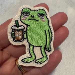 May include: Embroidered patch featuring a green frog with a tired expression, holding a brown iced coffee. The frog has pink cheeks and dark circles under its eyes. The patch has a white glittery border.