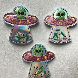May include: Three embroidered alien spaceship patches. Each patch features a green alien in a pink and yellow accented UFO. The UFOs have a holographic, iridescent finish and are filled with small, colorful shapes.