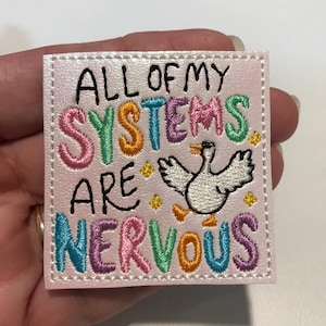 May include: A pink embroidered patch with the phrase "ALL OF MY SYSTEMS ARE NERVOUS" in colorful thread. A cartoon goose with outstretched wings is also embroidered on the patch. The patch is square with a white border.