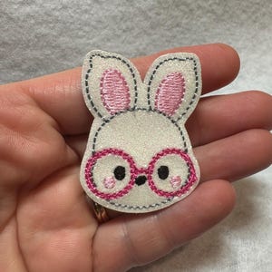 May include: A white bunny-shaped patch with pink inner ears and pink-framed glasses. The bunny has black eyes and is outlined with black stitching. The patch is held in a hand.