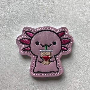 May include: Pink embroidered axolotl holding a brown cup with a red heart and a green straw. The axolotl has pink gills and black eyes.